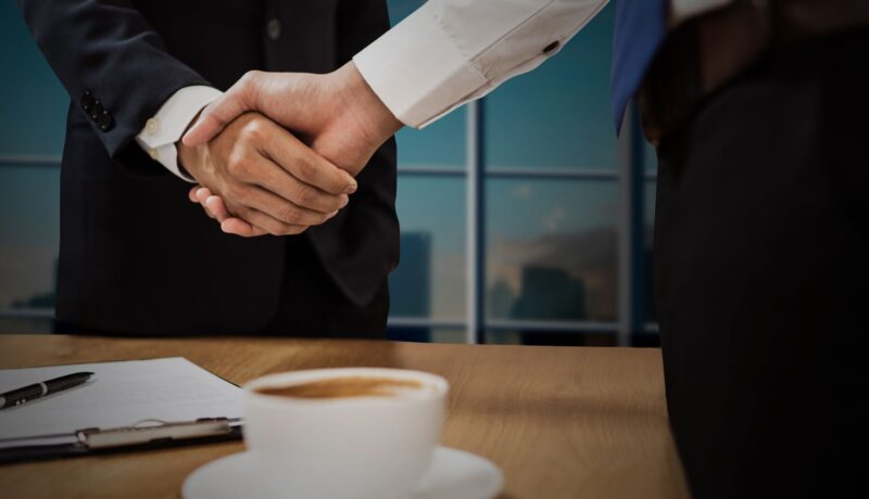 Two professionals shaking hands across a desk with cityscape in the background.