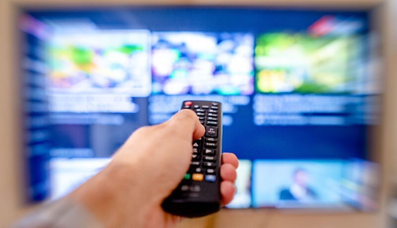 Hand holding remote control pointing at a blurred TV screen in a cozy indoor setting.