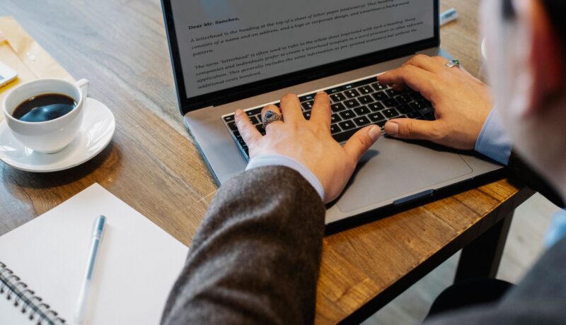 Crop faceless male entrepreneur in formal wear sitting at table with cup of coffee and notebook while working on project in laptop