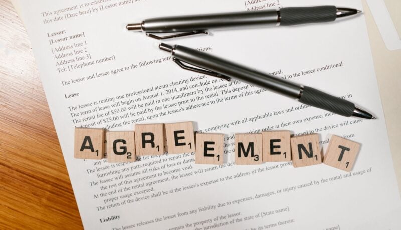 A document with Scrabble tiles spelling 'AGREEMENT' and two pens, symbolizing contract signing.
