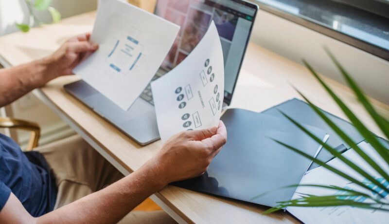 Crop anonymous male designer in casual wear sitting at table with opened laptop and folders and reading papers