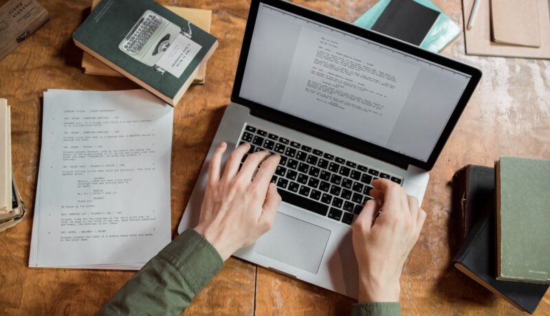 High-angle view of hands typing on a laptop surrounded by books and papers.