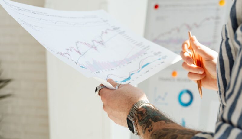 Close-up of a tattooed man's hands holding financial charts, with focus on data analysis and stock market trends.