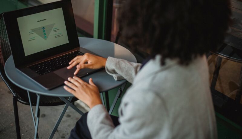 Back view of anonymous ethnic female in suit working with chart on computer while sitting at table in outdoor cafe during remote work